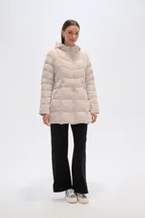 Campera acolchada color beige claro, de largo medio, con capucha y cierre frontal. Presenta costuras horizontales y diagonales que crean un efecto matelassé.