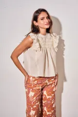 Blusa sin mangas de color crudo, con diseño oversize, cuello redondo y detalles de nido de abeja bordado en el pecho, acompañado de volados en los hombros.