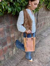 Matera de cuero con textura tipo crocco, combinando tonos rosado y marrón. Presenta asas cortas de cuero marrón y un detalle de borlas colgantes en el frente.