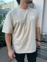 Remera de manga corta, corte relajado y color beige claro con efecto lavado. Presenta un estampado grande en la espalda en color blanco con el texto "Dropz Corp." sobre la silueta de un ángel en línea continua, junto a un código de barras y texto pequeño. En el frente lleva un logo sutil en el pecho.