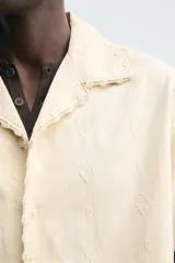Camisa de corte relajado confeccionada en tejido de algodón, con cuello bowling y manga corta. Presenta un diseño con bordados geométricos a tono y cierre frontal de botones.
