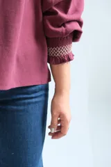 Blusa bordó de corte holgado, con escote en V y cuello fruncido. Presenta mangas tres cuartos con puños elásticos y detalles de volados.
