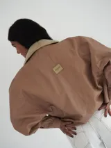 Campera oversize de cuero tostado con solapas y forro interno de corderito sintético azul marino, mangas anchas y detalles de costuras trenzadas.