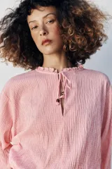 Blusa de manga larga con estampado de cuadros vichy en tonos rosado y blanco. Presenta cuello redondo con volante, cordón ajustable en el escote con apliques decorativos, y puños fruncidos con tiras para atar.