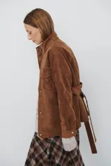 Chaqueta de gamuza en tono marrón oscuro, con cuello alto, cierre frontal con cremallera y botones a presión, bolsillos frontales con solapa y cinturón ajustable en la cintura.