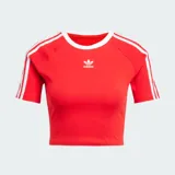 Remera corta roja ajustada al cuerpo, de manga corta, con cuello redondo blanco y las icónicas tres líneas blancas de Adidas en las mangas. Cuenta con el logo del trifolio bordado en el pecho.