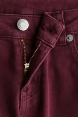 Pantalón de jean color bordó, de tiro bajo, corte ajustado hasta la rodilla y acampanado desde allí hacia abajo. Presenta cierre y botón frontal, bolsillos abiertos delanteros y bolsillos traseros con solapa y botón.