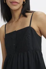 Vestido midi de color negro con tirantes finos, escote cuadrado y cuerpo con detalle de alforzas y botones frontales. Presenta una falda amplia con corte escalonado y bolsillos laterales.