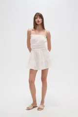 Vestido corto de algodón color blanco, con breteles finos tipo lencero, escote recto con detalle de volado y cintura con elástico fruncido. La falda presenta un diseño holgado con volado en el ruedo.