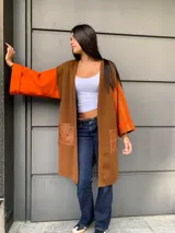 Saco largo de lana merino en color marrón, con diseño de bloques de color naranja en las mangas y detalles de bolsillos en cuero sintético. Presenta una manga con rayas naranjas y marrones en un lado y manga lisa naranja en el otro.