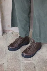 Borcego de cuero marrón con cordones jaspeados y suela track negra con costura blanca visible. Presenta un diseño con recortes en la puntera y laterales.