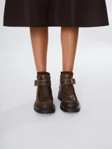 Bota corta tipo borcego de cuero marrón con suela track negra. Presenta costuras a la vista en la puntera y una tira ajustable con hebilla metálica en el tobillo.