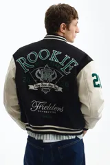 Campera estilo varsity con cuerpo negro y mangas en contraste color crema. Presenta múltiples parches y bordados decorativos en el frente y las mangas, cuello, puños y ruedo acanalados a rayas, y cierre frontal con botones a presión.
