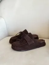 Zueco tipo mocasín de gamuza color marrón chocolate, con detalle de costuras visibles en el borde y cordones decorativos con borlas en el empeine.