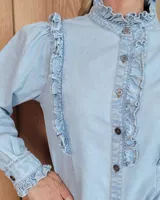Camisa de denim celeste con cuello alto con volados, botonadura frontal y detalles de volados verticales a lo largo del frente. Cuenta con mangas largas abullonadas y puños con volados.