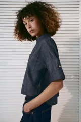 Camisa crop efecto denim color negro, con cuello solapa, escote en V, mangas cortas con trabilla y bolsillos frontales de parche. Cierre frontal con botones.