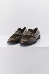 Mocasín color marrón claro, con capellada de cuero liso y aplique de gamuza en marrón oscuro. Tiene punta redondeada y suela plana de goma negra.