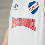 Musculosa de básquetbol Umbro del Club Nacional de Football, color blanco con detalles en azul y rojo en cuello y sisas. Presenta el logo de Umbro y el escudo del club en el frente, junto con la inscripción Nacional en letras rojas con borde azul.