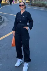 Mono de gabardina negro con cuello camisero, cierre frontal con botones metálicos, mangas largas y bolsillos tipo cargo en el pecho y las piernas. Incluye un cinturón ancho con hebilla del mismo tono para ajustar la cintura.