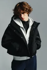 Anorak corto de color negro con acolchado interior, cuello subido y capucha. Presenta mangas largas con puños elásticos, bolsillos delanteros con cierre y bajo ajustable con cordones. Cierre frontal con cremallera termosellada y botones a presión.