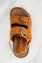 Sandalia de gamuza color tostado con base de goma marrón, diseño de tiras anchas con hebillas metálicas doradas y ajuste en el talón.