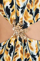 Trikini de una sola pieza con estampado de hojas en tonos mostaza, negro y blanco. Presenta un diseño asimétrico con un solo hombro y recortes laterales unidos por un adorno metálico dorado con forma de hoja.