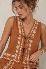 Conjunto de dos piezas color tostado con bordados florales en hilo blanco, compuesto por una blusa sin mangas con escote en V y cierre frontal con lazos, y un short tiro alto.