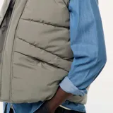 Chaleco acolchado puffer color gris, sin mangas, con cuello alto, cierre frontal de cremallera con solapa de botones a presión y bolsillos laterales amplios.