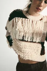 Jersey de punto con cuello redondo y manga larga, caracterizado por un diseño de parches en tonos tierra y crema. Presenta una textura variada con detalles de flecos y tejido en relieve.