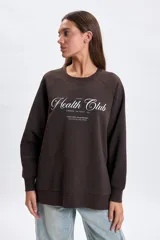 Buzo deportivo de corte oversize en color marrón, con cuello redondo y mangas largas tipo raglán. Presenta una estampa frontal con la inscripción 'Health Club' en tipografía cursiva y detalles de texto en blanco.