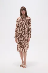 Vestido tipo camisa de corte holgado, con cuello clásico, cierre frontal de botones y mangas largas. Presenta un estampado botánico en tonos marrones y beige.