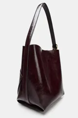 Bolso tipo saca de diseño asimétrico con acabado brillante, asa ajustable con hebilla lateral y cierre mediante botón imán.