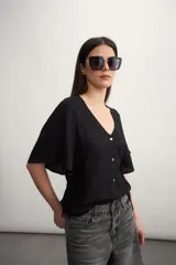 Camisa negra con botones dorados, escote en V y mangas cortas amplias con lazos laterales. 