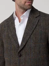 Blazer de lana Harris Tweed con patrón de espiga y cuadros sutiles en tonos tierra, azul y naranja. Presenta cierre frontal de dos botones, solapas clásicas y bolsillos con solapa. Incluye forro interior satinado.