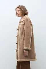 Abrigo de estilo duffle coat en simil gamuza color tostado, con interior y bordes de piel sintética al tono. Presenta cierre frontal mediante alamares de cuero sintético y madera, cuello solapa y mangas largas.