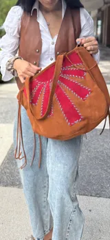 Cartera tipo tote bag de gamuza color marrón, con diseño reversible. Presenta un aplique de gamuza roja con costuras decorativas en hilo azul en una de sus caras, mientras que la otra cara es lisa. Incluye tiras laterales decorativas.