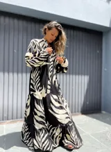 Vestido largo tipo túnica con estampado de hojas en contraste blanco y negro. Presenta cuello camisero con botones, mangas largas amplias y falda con volados escalonados.