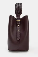Bolso tipo saca de color bordó, con asa de mano trenzada y cierre mediante botón imantado. Incluye bolsillo interior con cremallera.