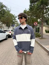 Buzo tipo polo de corte relajado y fit boxy, con diseño de bloques de color en gris y blanco. Presenta cuello camisero, cierre frontal parcial y logo bordado en el pecho.