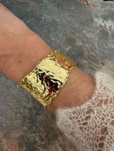 Pulsera rígida tipo brazalete de diseño ancho y textura irregular con acabado dorado brillante.