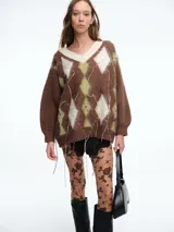 Sweater de tejido de punto con diseño de rombos (argyle) en tonos marrón, verde oliva y crema. Presenta escote en V, mangas largas con puños ajustados y detalles de hilos colgantes decorativos en el frente que le otorgan un aspecto desgastado y moderno.