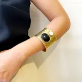 Brazalete ancho de bronce con acabado martillado, que presenta dos piedras circulares engarzadas: una de nácar blanco de gran tamaño y otra de piedra natural oscura de menor tamaño. Es una pieza de diseño ajustable tipo cuff.
