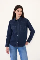 Camisa de jean de corte clásico estilo western, confeccionada en denim azul oscuro con pespuntes contrastantes. Presenta cuello camisero, bolsillos frontales con solapa y cierre de botones metálicos a presión.