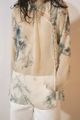 Camisa de cuello redondo y manga larga con diseño de estampado artístico en tonos azules sobre fondo beige. Presenta un cierre frontal asimétrico con botones forrados a tono y una abertura lateral en el escote.