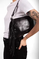 Cartera bandolera de cuero negro con flecos colgantes en el lateral, cierre superior y correa ajustable con argollas metálicas.