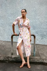 Vestido midi de corte camisero con estampado floral multicolor sobre fondo rosa claro. Presenta cuello solapa, manga corta, cierre frontal con botones y cinturón del mismo tejido con hebilla forrada.