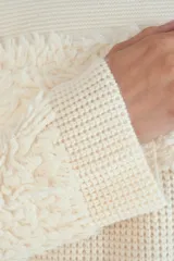 Suéter de punto color crema con diseño de paneles texturizados tipo peluche en el pecho y en los puños. Presenta cuello redondo y corte holgado.
