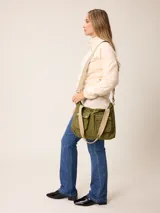 Bolso tipo bandolera de lona color mostaza, con dos bolsillos frontales con solapa y cierre de broche a presión. Cuenta con correas de hombro negras y una correa larga ajustable y extraíble. Presenta herrajes metálicos en tono bronce.