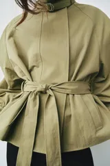 Cazadora de color verde militar, con cuello alto, manga larga abullonada con puños y cierre frontal con botones a presión ocultos. Incluye un cinturón ajustable del mismo tejido para entallar la cintura y bolsillos delanteros de vivo.