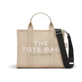 Bolso tipo tote de lona en color beige, con estampado frontal en blanco y gris que dice 'THE TOTE BAG MARC JACOBS'. Cuenta con doble asa superior, cierre de cremallera y correa larga ajustable y extraíble en color negro.